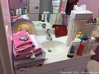 Photo showing bathroom counter with towels, toiletries, decorative hat, basket with tissue box, and toiletry bottles.