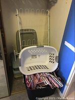 White metal rack with flat plastic storage totes stacked underneath, white plastic laundry hamper placed on top. Larger plastic tote visible at base containing hangers and reusable bags.