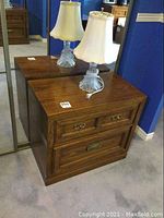 Wood night stand with two drawers seen from front, showing wood grain and brass-tone drawer handles. Includes 17 inch glass lamp with beige lampshade on top.