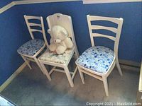 Photo showing the pair of painted wood chairs with blue patterned cushioned seats and the additional chair with floral cushioned seat and teddy bear, placed against a blue wall