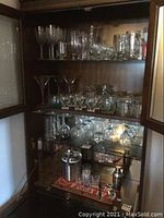 Glassware of various types arranged on glass shelves inside wooden cabinet with mirror backs, showing wine glasses, martini glasses, and other drink glasses.
