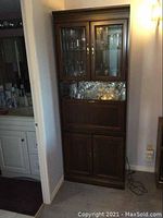 Front view of a tall wooden bar cabinet with glass doors on top showing glassware inside.
