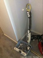 Shark steam mop leaning against wall with plastic bin containing mop pads and attachments on floor next to it.