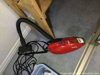 Red Dirt Devil Scorpion handheld vacuum cleaner with flexible hose attachment on carpeted floor near wooden furniture.