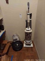 View of black iRobot Roomba Pet Series robotic vacuum next to white Shark upright vacuum in a corner. Both vacuums and accessories shown clearly.