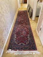 Full length view of the rug runner laid in a hallway showing its red and blue patterned design with cream and dark blue borders and white fringes at one end.
