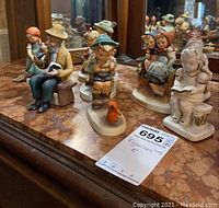 Figurines displayed on marble surface showing children reading, holding birds, and sitting. Tag with lot number and measurement.