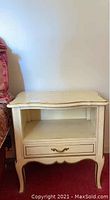 Front view of one cream colored nightstand showing drawer, open shelf, curved apron, and brass handle.