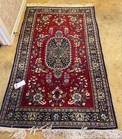 Full view of a red floral area rug with navy blue border and white fringe, placed on a beige carpeted floor.