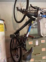 Side view of black and blue Nomad bicycle with 28-inch wheels propped vertically in garage environment, showing the front suspension fork, pedals, and handlebars.