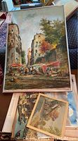 Framed painting showing a European street scene with buildings on each side, market stalls with red umbrellas, and figures of people. It is signed by J. Vando in the lower corner.