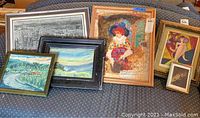 Six framed art pieces arranged on a couch viewed from front angle showing variety of styles and frame colors