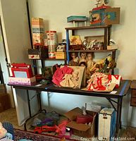 Folding table with dolls and various items including boxes and fabric items on top and underneath.