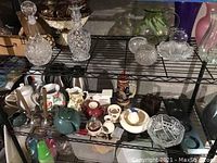 Wide view of three shelves containing assorted decorative items including crystal decanters, pitchers, bowls, and various containers.