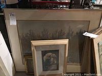 Three framed wall art pieces stacked against a wall; largest is a landscape print with trees in muted colors, medium is an architectural print with an archway, the smallest is an additional framed print.