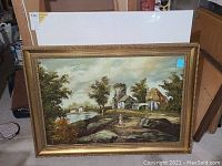 Framed landscape oil painting with rustic rural scene, showing trees, water, rocky path, buildings including stone tower and thatched roof house, with a person in pink dress.