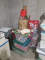 Stack of plastic bins and cardboard boxes containing various Christmas decorations including tree ornaments and holiday decor.
