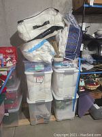 Stack of three white/cream colored bedding items in clear storage bags on top and four white plastic storage totes below with gray lids and some visible fabric inside.