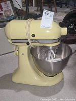 Side angle of the yellow KitchenAid mixer showing the power cord, control knob, and stainless steel bowl attached.