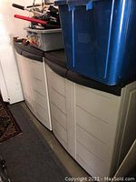 Two white plastic storage cabinets side by side with black tops, measuring about 30x19x36 inches each. Items on tops not included.