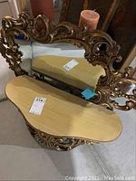 Front view of ornate gold-toned mirror with matching wall shelf below, candle on the shelf.