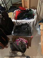 Photo of a white laundry basket filled with assorted bags, including black, red, and green soft bags, with two additional bags in front on the floor.