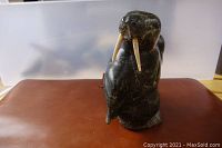 Dark stone Inuit carving of a walrus with detailed tusks and realistic sculptural features, standing 7 inches tall on a flat surface with plain backdrop.