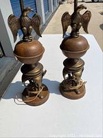 Pair of metal lamps with eagle-shaped finials on top, showing wear and cords
