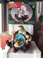 Santa figurine sitting holding a snow globe with box in background showing same design