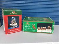Boxed 15-inch Kurt S. Adler musical snow blowing fiber optic tree and Holly Dearies figurine set boxes shown side by side.