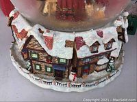Close-up of the base of a snow globe showing detailed houses with snow-covered roofs, brick walls, and a snowman figure, depicting a Christmas village scene