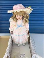 Doll standing in original box, wearing pink lace dress, blue ribbon, floral hat, and white shoes