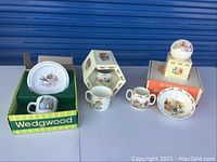 Overall view of Bunnykins and Wedgwood children's tableware items displayed on a surface, showing multiple pieces including cups, bowls, and a decorative ornament, some in original boxes.