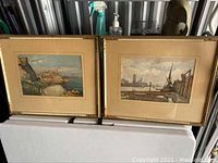 Two framed watercolor prints resting side by side, showing coastal castle and harbor scenes in gold-toned frames with beige mats.