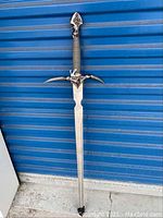 Full length image of the replica Excalibur sword leaning against a blue wall, showing ornate guard, wrapped handle, and full blade.