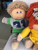 Photo showing two vintage Cabbage Patch Kids dolls, one with curly brown yarn hair in green and gray outfit numbered 31, adjacent to one with orange yarn hair in yellow dress and sequin crown.