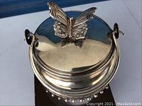 Top view of silver-toned round tea caddy with a butterfly-shaped handle on lid, showing its polished surface and carry handle.