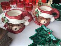 Photo of two bear-shaped ceramic mugs with Santa hats placed on a table with other Christmas-themed items including red glasses and green leaf-shaped dishware.