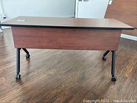 Front view of a rolling office desk showing the red/brown wood veneer finish and black metal legs with casters.