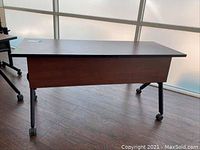 Front and side view of rolling office desk showing wood veneer surface, black legs, and caster wheels.