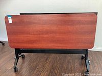 Front view of rectangular rolling desk with red/brown wood veneer top, black frame, and caster wheels.