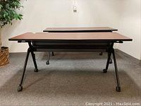Front angle view of rolling wood veneer desk with black metal frame on wheels, placed on carpet with wall and plant in background
