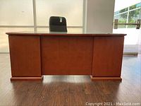Front view of the large wooden desk with a glass top, showing its full width and wood grain finish, on a dark wood laminate floor.