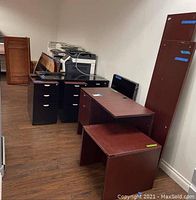 Overview of the office furniture grouping including two metal filing cabinets with multiple drawers, multiple wooden desks and panels stacked and arranged together in a room with wood laminate flooring.