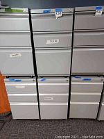 Front view of multiple gray metal two-drawer filing cabinets in a row, labeled for desks, showing uniform design and metallic handles.