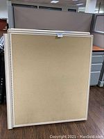 Front view of three beige fabric office cubicle partitions with white metal frames stacked together inside an office environment.