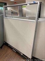 Photo showing a metal-framed office cubicle partition with fabric lower panel and upper glass panel, standing upright in an office environment.