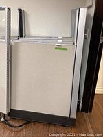 Close-up photo showing single cubicle partition panel with light gray fabric, silver metal frame and black base piece, placed upright on floor.