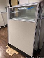 Single office cubicle partition panel with metal frame, fabric lower panel and upper glass section. Panel is upright, resting on black base supports.