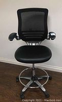 Front view of black adjustable office chair showing mesh backrest, padded seat, armrests, footring, and caster wheels on wood floor.